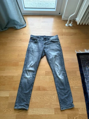 G star raw  Gråa jeans  - Säljer ett par gråa jeans med klassisk femficksdesign och dragkedja. Jeansen har en snygg tvättad look och raka ben. Perfekta för dig som gillar en stilren och modern vibe. Materialet är denim, vilket ger både komfort och hållbarhet. Storlek 32/34. Regular tapered fit.