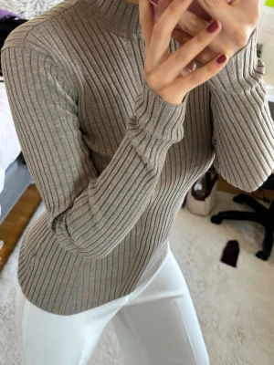 Beige ribbstickad polotröja - Säljer en beige ribbstickad tröja med hög hals och långa ärmar. Tröjan har en figurnära passform och är tillverkad i ett mjukt, stretchigt material som sitter snyggt på kroppen. Perfekt att matcha med jeans eller kjol. HM