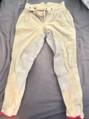 Beige ridbyxor med förstärkning - Snygga beige ridbyxor med grå förstärkning på insidan av benen och röd kant längst ner. Byxorna har hög midja, bälteshällor och knäppning fram. Perfekta för ridning tack vare det slitstarka materialet och den bekväma passformen.