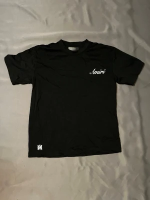 Svart Amiri t-shirt  - Snygg svart t-shirt från Amiri med vit logga på bröstet och en liten detalj nertill. Klassisk rund hals och korta ärmar. Perfekt basplagg med stilren design och skön passform.