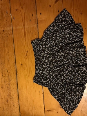 Svart blommig volangkjol - Söt svart kjol med vitt blommönster och volangkant. Kjolen har en asymmetrisk form och är kort i modellen, perfekt för en trendig look. Materialet känns lätt och flowy, vilket ger en skön känsla.
