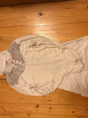 Vit blus med spetsdetaljer - Stilren vit blus med lång ärm och hög krage. Blusen har vackra spetsdetaljer över axlar och bröst samt knappar framtill. Perfekt för en chic och elegant look. Materialet känns mjukt och lätt mot huden.