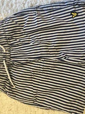 Randiga badbyxor från Lyle & Scott - Snygga badbyxor från Lyle & Scott med marinblå och vita ränder samt gul logga på sidan. De har resårmidja med vit dragsko och meshfoder på insidan för extra komfort. Perfekta för strand eller poolhäng. Storlek 8-9