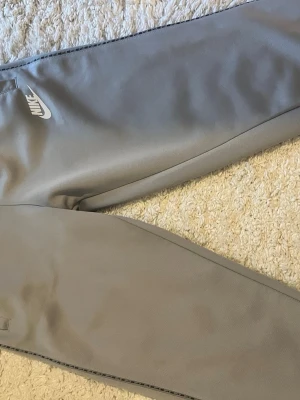Gråa mjukisbyxor från Nike - Snygga gråa mjukisbyxor från Nike med vit logga på vänster lår. Byxorna har en smal passform och resår i midjan för extra komfort. Perfekta för chill dagar eller träning. Tillverkade i mjukt och bekvämt material.