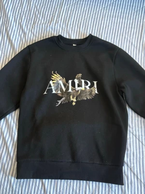 Svart sweatshirt från Amiri med örn - Cool svart sweatshirt från Amiri med stort tryck av en örn och AMIRI-logga på bröstet. Tröjan har rund halsringning, ribbade muddar och är gjord i mjukt material. Perfekt för dig som gillar streetwear och statement-plagg. Passar för barn storlek 158-164 