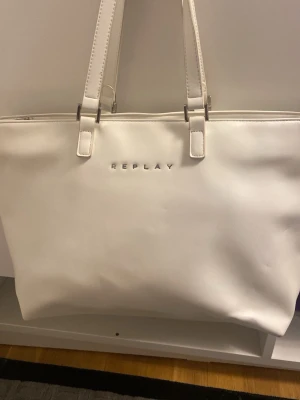 Vit handväska från Replay - Stilren vit handväska från Replay med två långa axelband och diskret logga framtill. Väskan har en klassisk rektangulär form och är tillverkad i slätt konstläder. Perfekt för dig som gillar minimalistisk och modern design.