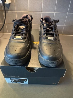 Nike Air Force 1 Low helsvarta sneakers - Säljer ett par klassiska Nike Air Force 1 Low sneakers i helsvart läder. Skorna har perforerad tå, svarta skosnören och metallbricka med 'AF1' på snörningen. Perfekt för dig som gillar stilrena och tidlösa sneakers.