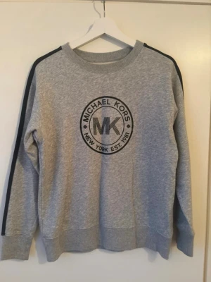 Grå sweatshirt från Michael Kors - Snygg grå sweatshirt från Michael Kors med svart logga och text på bröstet. Tröjan har svarta ränder längs ärmarna och ribbade muddar vid hals, ärmslut och nederkant. Tillverkad i mjukt bomullsmaterial för en bekväm känsla.