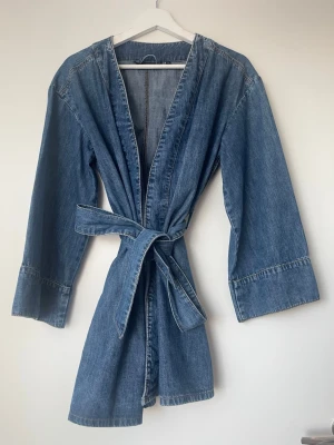 Jeanskimono med knytskärp - Snygg jeanskimono från Keep A i klassisk blå denim. Modellen har vida ärmar, öppen front och ett matchande knytskärp i midjan. Perfekt för dig som vill ha en avslappnad men trendig look. Materialet är mjukt denimtyg som ger en cool vibe.