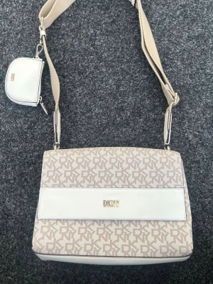 Beige axelremsväska från DKNY - Snygg beige axelremsväska från DKNY med monogrammönster och vita detaljer. Väskan har justerbar bred axelrem och ett litet matchande myntfodral. Guldigt DKNY-emblem framtill och stilren rektangulär form. Perfekt för dig som gillar trendiga accessoarer.