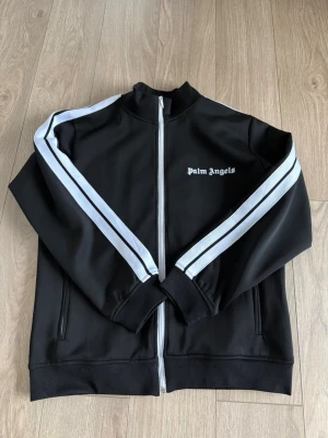 Palm Angels svart track suit - Svart track jacket från Palm Angels med vita ränder längs ärmarna och vit dragkedja. Jackan har hög krage, ribbade muddar och logga på bröstet. Tillverkad i ett glansigt, sportigt material som passar perfekt till streetwear och träning. Matchande byxor i samma stil. 