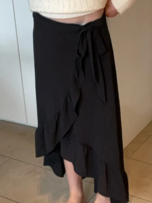 Svart omlottkjol med volang - Svart långkjol i omlottmodell med volangkant och knyt i midjan. Kjolen har en snygg asymmetrisk form och faller mjukt längs benen. Perfekt för dig som gillar en stilren men ändå trendig look.