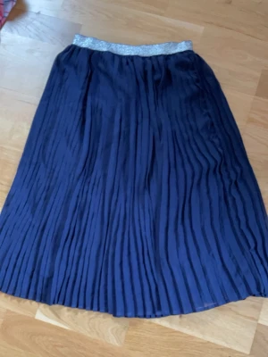 Mörkblå plisserad långkjol - Snygg mörkblå plisserad långkjol med glittrigt silverresår i midjan. Kjolen har ett elegant fall och passar perfekt till både vardagslooken och mer uppklädda tillfällen. Materialet känns lätt och följsamt.