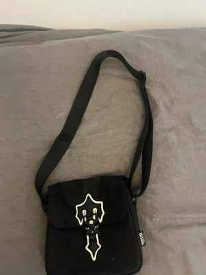 Svart axelväska från TRAPSTAR - Snygg svart axelväska från TRAPSTAR med justerbar rem och unik vit detalj på locket. Väskan har fyrkantig form och stängs med spänne framtill. Tillverkad i slitstarkt tygmaterial och passar perfekt för dig som vill ha en cool och praktisk väska inför sommaren