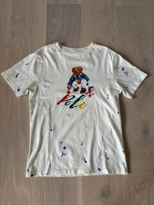 Vit Polo Ralph Lauren t-shirt björn - Vit t-shirt från Polo Ralph Lauren med färgstänk och en stor tryckt björn på framsidan som målar ordet 'Polo' i rött, blått, grönt och gult. Klassisk passform och korta ärmar. Perfekt för dig som gillar lekfulla och unika prints. Vid intresse är det bara att höra av sig. Pris går att diskutera 