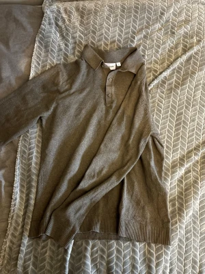 Olivgrön pikétröja från Kappahl - Snygg olivgrön pikétröja från Kappahl med klassisk krage och tre knappar framtill. Tröjan har långa ärmar och ribbade muddar vid ärmslut och nederkant. Perfekt för en stilren och avslappnad look.