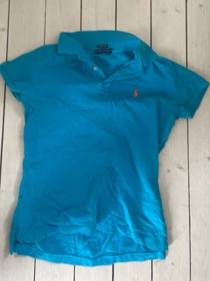 Blå pikétröja från Ralph Lauren - Klassisk blå pikétröja från Ralph Lauren med korta ärmar och krage. Tröjan har knappar vid halsen och den ikoniska lilla loggan broderad i orange på bröstet. Perfekt till jeans eller shorts för en avslappnad look.