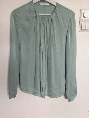 Ljusgrön blus från BOSS - Stilren ljusgrön blus från BOSS med knappar framtill och lätt rynkad detalj vid halsen. Tillverkad i ett tunt, mjukt tyg som ger ett elegant fall. Perfekt till jeans eller kjol för en clean look.