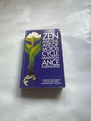 Zen and the Art of Motorcycle Maintenance - En klassisk roman på engelska av Robert M. Pirsig med blått omslag. Boken har ett unikt omslagsmotiv med en vit blomma och en skiftnyckel. Perfekt för dig som gillar filosofiska resor och reflektioner kring livet.