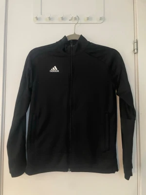 Svart träningsjacka från Adidas - Säljer en svart träningsjacka från Adidas med dragkedja framtill och vit Adidas-logga på bröstet. Jackan har hög krage, långa ärmar och två sidofickor. Perfekt för träning eller uppvärmning. Tillverkad i ett lätt och funktionellt material.