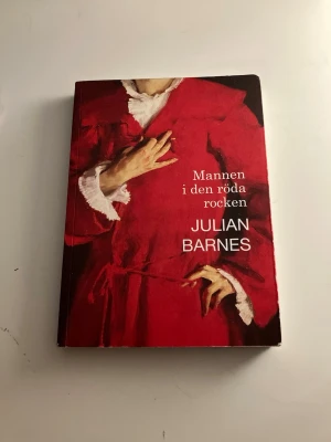 Mannen i den röda rocken - Romanen 'Mannen i den röda rocken' av Julian Barnes har ett omslag i rött med en målning av en person i röd rock. Boken är tryckt på papper och har mjuka pärmar. Texten på omslaget är vit och boken är på svenska.