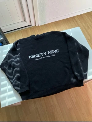Svart stickad tröja NINETY NINE - Svart stickad tröja med texten 'NINETY NINE' och mindre text under på ryggen. Tröjan har rund halsringning, ribbade muddar vid ärmslut och nederkant. Perfekt för dig som gillar streetwear och enkel stil.
