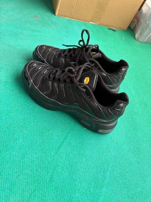 Svarta Nike Air Max Plus sneakers - Nike Air Max Plus sneakers i helsvart med diskreta grå detaljer och mesh-överdel. Skorna har en ikonisk vågig design, synlig Air Max-dämpning i sulan och gula detaljer på plösen. Perfekta för dig som gillar streetwear och sportig stil.