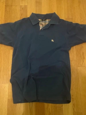 Blå pikétröja från Burberry - Klassisk blå pikétröja från Burberry med korta ärmar och den ikoniska rutiga detaljen i kragen. Liten broderad logga på bröstet. Perfekt till jeans eller shorts för en clean och stilren look. Har dock lite fläckar men inget man märker bara sådär de står dock att de är xxl men den sitter som s/m