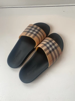 Burberry sandaler med rutigt band - Säljer ett par snygga sandaler från Burberry med klassiskt beige, svart och rött rutigt band över foten. Sulan är svart och har Burberry-loggan präglad. Perfekta för sommaren och riktigt trendiga till streetwear-looken.