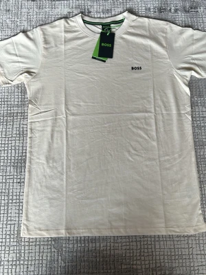 Vit t-shirt från BOSS - Snygg vit t-shirt från BOSS med klassisk rund hals och diskret BOSS-logga broderad på bröstet. Tillverkad i mjukt bomullsmaterial som känns skönt mot huden. Perfekt basplagg för en clean och stilren look.