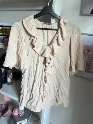 Beige volangblus från Carin Wester - Supersöt beige blus från Carin Wester med volangdetaljer längs ringningen och framtill. Blusen har korta ärmar och passar perfekt till ett par vita jeans. Tillverkad i ett mjukt, lätt tyg som ger en avslappnad men ändå chic look.