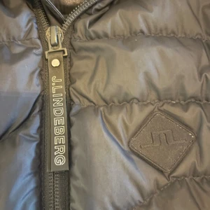 Svart dunjacka från J.Lindeberg - Snygg svart dunjacka från J.Lindeberg med quiltad design och dragkedja framtill. Jackan har en diskret logotyp på bröstet och J.Lindeberg-tryck på dragkedjan. 