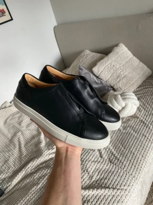 Myrqvist Alnö Lacelese lädersneakers - Nypris 2200kr Stilrena svarta sneakers i slätt och lätt strukturerat läder med vit, tjock sula. Skorna har en minimalistisk design utan synliga snören och en låg profil. Insidan är ljusbeige vilket ger en snygg kontrast. Perfekta för dig som gillar clean och modern stil.