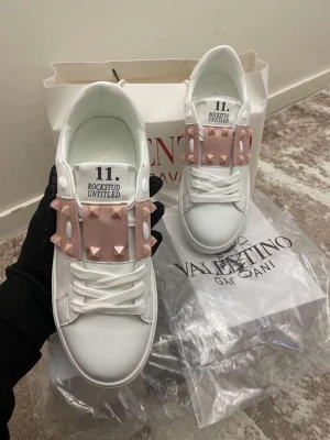 Valentino Rockstud Untitled sneakers - Snygga vita sneakers från Valentino med ljusrosa detaljer och ikoniska pyramidnitar på ovandelen. Modellen heter Rockstud Untitled och har en chunky sula, vita skosnören och coola detaljer som sticker ut. Perfekta för dig som vill ha något unikt och trendigt.