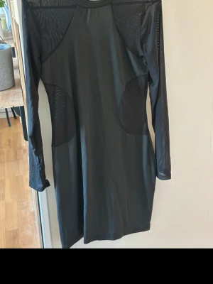 Svart meshklänning med lång ärm - Svart klänning i meshmaterial med lång ärm och rund hals. Klänningen har transparenta meshpartier på ärmar och axlar samt en stilren, kroppsnära passform. Perfekt för dig som gillar en edgy och modern look.