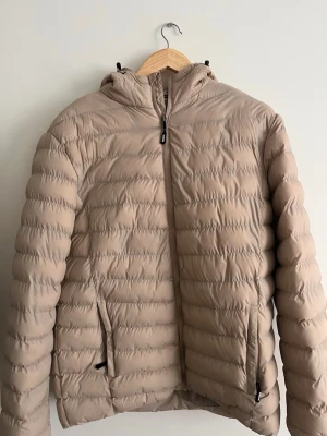  pufferjacka  - Säljer en beige vindjacka med huva och dragkedja framtill. Jackan har två sidofickor med dragkedja och quiltad design som ger en puffig känsla. Tillverkad i syntetmaterial och lätt vadderad för extra komfort. Perfekt för dig som vill ha en stilren och funktionell jacka.