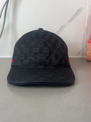 Svart GGi keps med GG-mönster - Säljer en svart keps från GG med det klassiska GG-mönstret över hela kepsen. På sidan finns en grön och röd rand som ger en ikonisk touch. Kepsen har böjd skärm och är tillverkad i ett slitstarkt tygmaterial. Perfekt för dig som vill ha en stilren och lyxig accessoar.