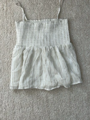 Vit smockad linnetopp med glitter - Supersöt vit linnetopp med smockad överdel och avtagbara tunna axelband. Toppen har ett skirt, glittrigt tyg med subtila silverränder och volangig nederdel. Perfekt för sommarens alla tillfällen och lätt att styla till jeans eller kjol. 