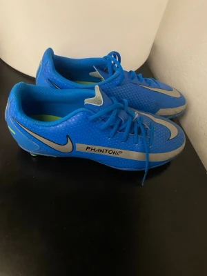 Nike Phantom GT2 blå fotbollsskor - Säljer ett par Nike Phantom GT2 fotbollsskor i blått med silverfärgade detaljer och stora Nike-loggor på sidorna. Skorna har snörning och är tillverkade i syntetmaterial för bra grepp och kontroll på planen. Perfekta för dig som vill sticka ut på fotbollsplanen.