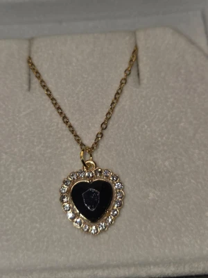 Halsband med hjärthänge och stenar - Guldigt halsband i rostfritt stål med ett hjärtformat hänge. Hänget har en svart mitt och är kantat av gnistrande klara stenar som ger en lyxig känsla. Kedjan är tunn och elegant, perfekt för att lyfta vilken outfit som helst. Kedjan är 45 cm + 5 cm förlängningskedja.