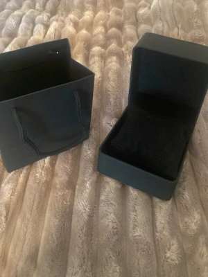 Svart presentpåse och ask - Säljer en stilren svart presentpåse med matchande ask, perfekt för smycken eller små accessoarer. Asken har mjuk insida och enkel design, medan påsen har korta handtag i tyg. Båda är i svart färg och passar till att ge bort presenter.