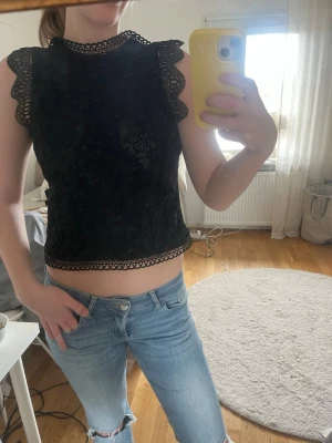 Svart spetsblus med volangdetaljer - Säljer en svart blus med vackra spetsdetaljer och volang längs ärmlösa axlar. Blusen har hög hals med dekorativ kant och är lite croppad. 