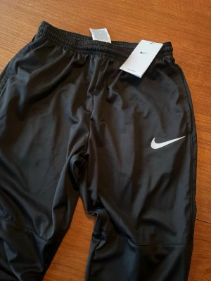 Svarta Nike Dri-FIT träningsbyxor - Säljer ett par svarta träningsbyxor från Nike med Dri-FIT-material som transporterar bort fukt. Byxorna har elastisk midja och den klassiska vita Nike-loggan på benet. Perfekta för gymmet eller löpning.