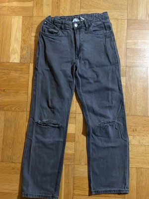 Gråa raka jeans med slitningar - Säljer ett par gråa jeans med raka ben och slitningar vid knäna. Jeansen har klassisk femficksdesign, knappgylf och en avslappnad passform. Perfekta för en cool och avslappnad stil.