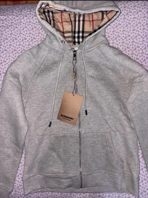Grå zip hoodie från Burberry - Snygg grå zip hoodie från Burberry med klassiskt rutigt foder i huvan. Hoodien har dragsko i huvan, fickor framtill och diskret Burberry-logga på bröstet. Tillverkad i mjuk bomullsblandning för skön känsla. Perfekt för en clean och stilren look.