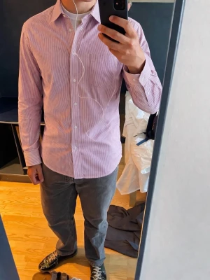 Rosa Randig skjorta från Acne Studios - Säljer en snygg randig skjorta från Acne Studios i ljusrosa och vit. Skjortan har klassisk krage, knappar framtill och långa ärmar. Tillverkad i en mix av linne och bomull för en luftig känsla. Perfekt för dig som gillar stilrena plagg med modern touch.