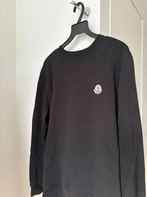Svart sweatshirt från Moncler - Svart sweatshirt från Moncler med rund halsringning och Moncler-logga broderad på bröstet. Tröjan har en klassisk passform och är tillverkad i mjukt material, perfekt för en clean och stilren look. Hör gärna av dig om du har några funderingar!