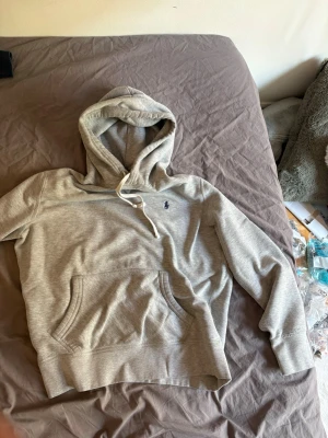 Grå hoodie från Polo Ralph Lauren - Snygg grå hoodie från Polo Ralph Lauren med klassisk marinblå logga broderad på bröstet. Hoodien har huva med vita dragsnören och en stor magficka framtill. Tillverkad i mjukt bomullsmaterial som är skönt mot huden. Perfekt för en avslappnad och stilren look.