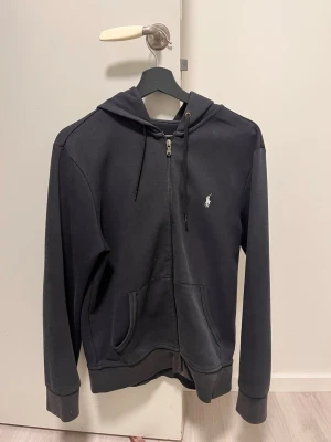 Svart zip hoodie från Polo Ralph Lauren - Snygg svart hoodie med dragkedja från Polo Ralph Lauren. Klassisk design med huva och dragsnören, två fickor framtill.