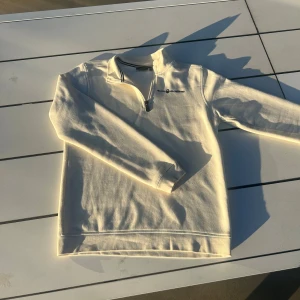 Vit Sail Racing half zip tröja - Säljer en stilren vit långärmad tröja från Sail Racing med halv dragkedja och broderad logga på bröstet. Tröjan har en klassisk passform och är tillverkad i mjuk bomull. Snygga detaljer vid kragen och snygg logga i nacken!